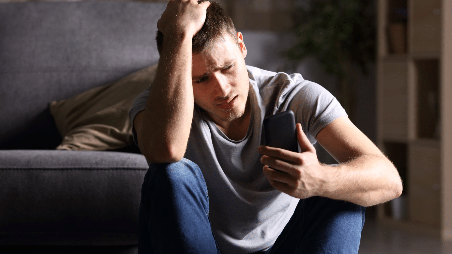 A young man sitting on the floor beside a sofa holding his phone and clutching his head while looking stressed at the screen