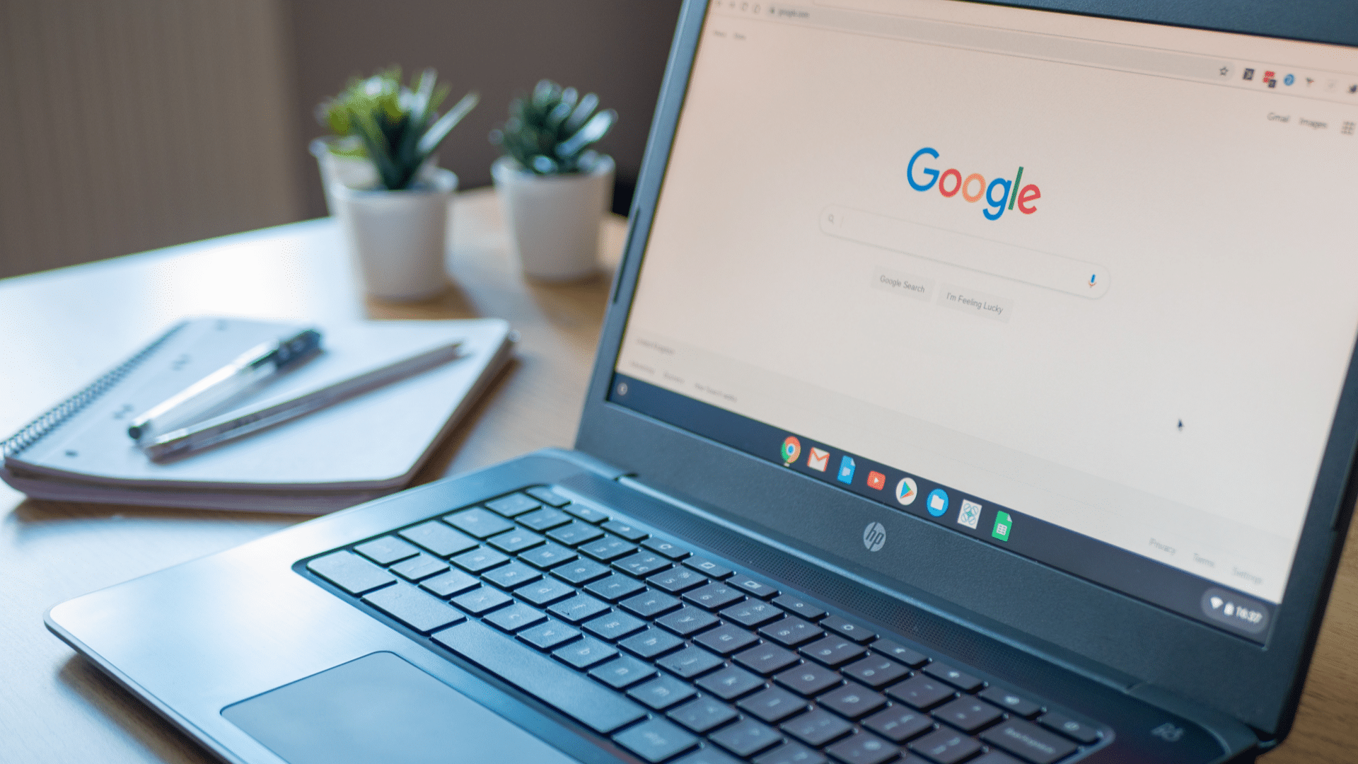 a chromebook on a desk displaying the google search homepage with notebooks and plants nearby 