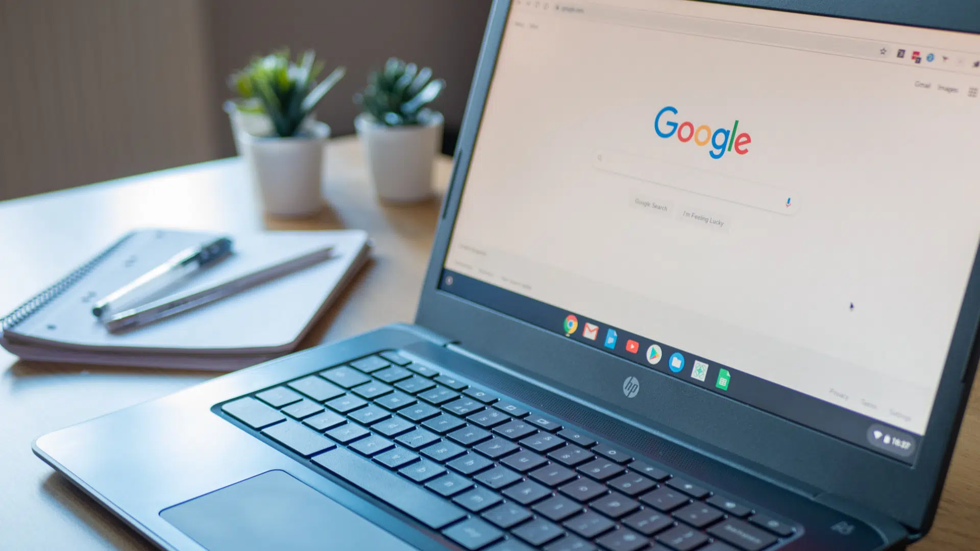 a chromebook on a desk displaying the google search homepage with notebooks and plants nearby 