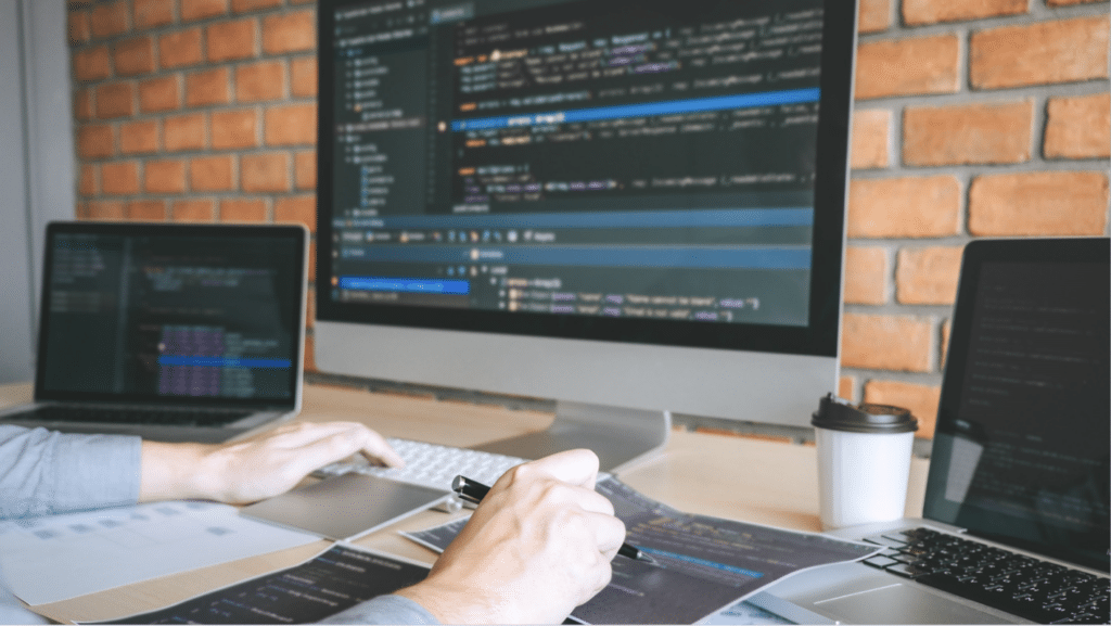 a cybersecurity person reviews and edits code on multiple screens, with a laptop, desktop, and papers with code printed out