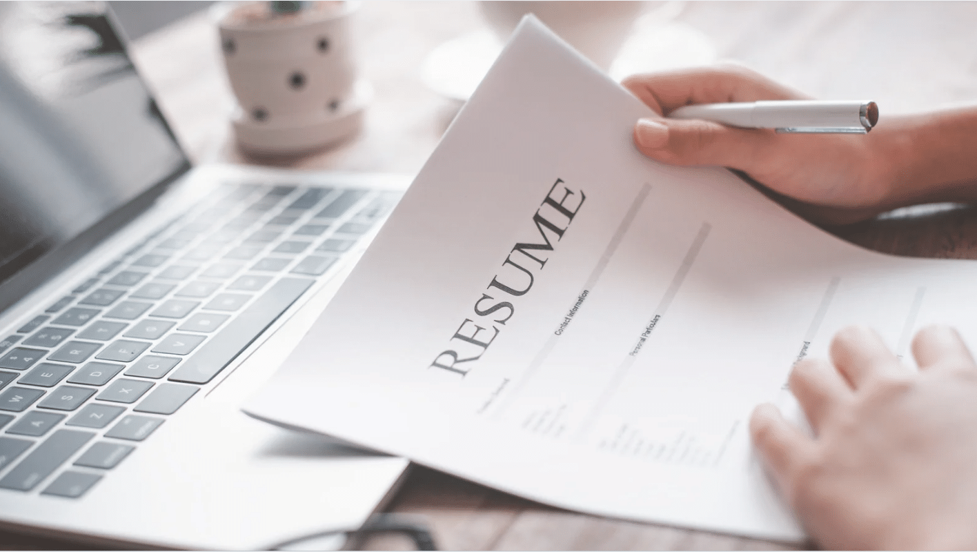 a person reviewing a printed cybersecurity resume with a pen in hand, sitting next to a laptop and a small plant on a desk.