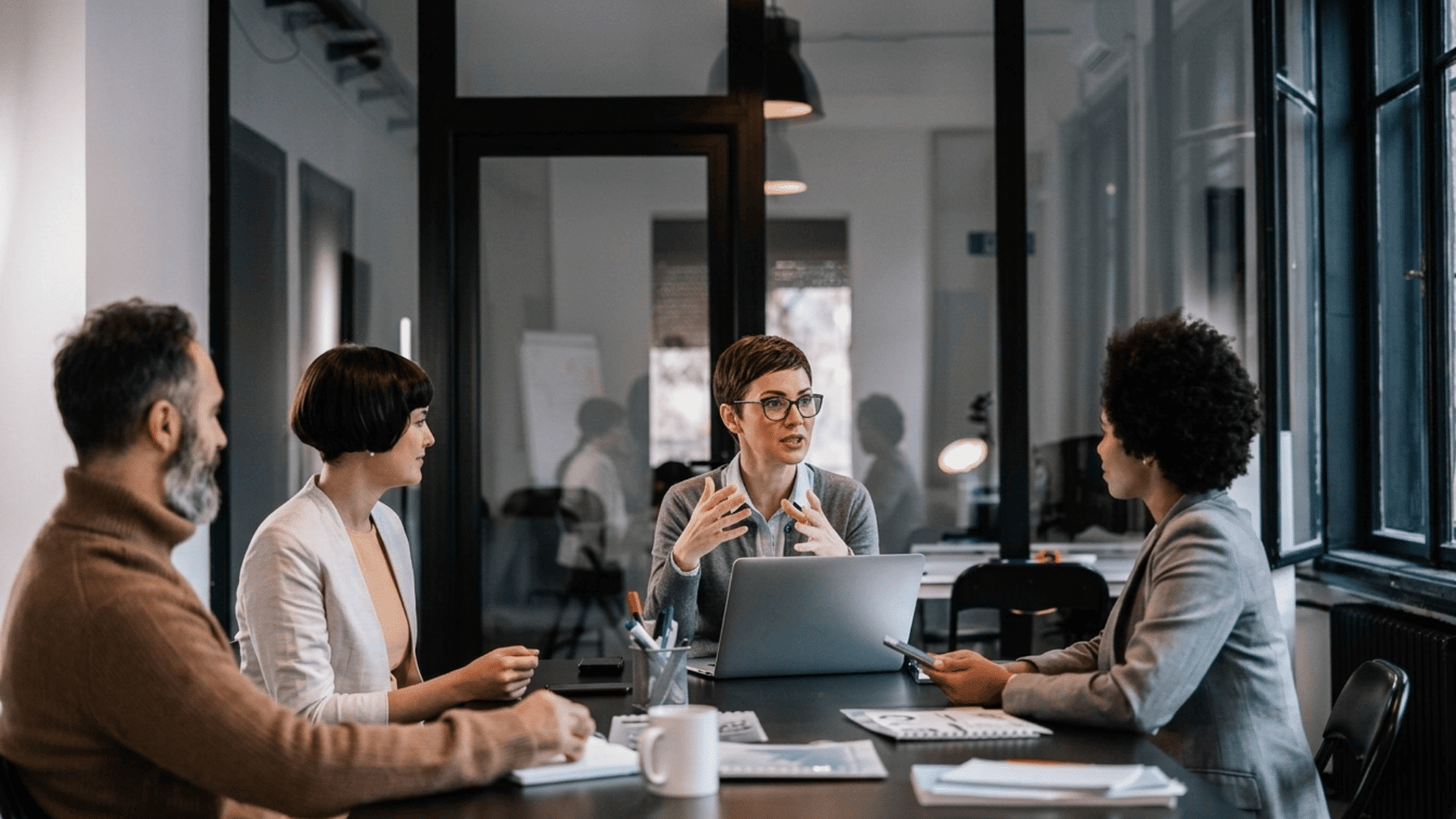 team meeting in modern office with four professionals discussing ideas around a laptop and sharing notes together