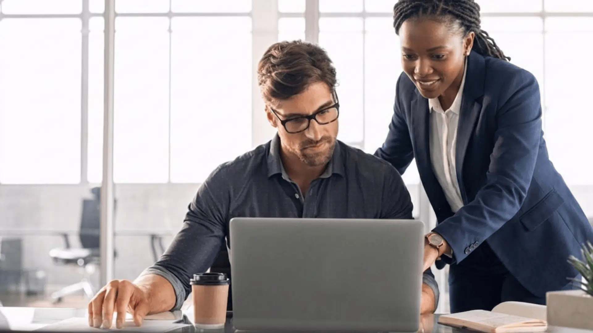 two professionals working together on a laptop in a bright office one explaining while the other focuses on the screen