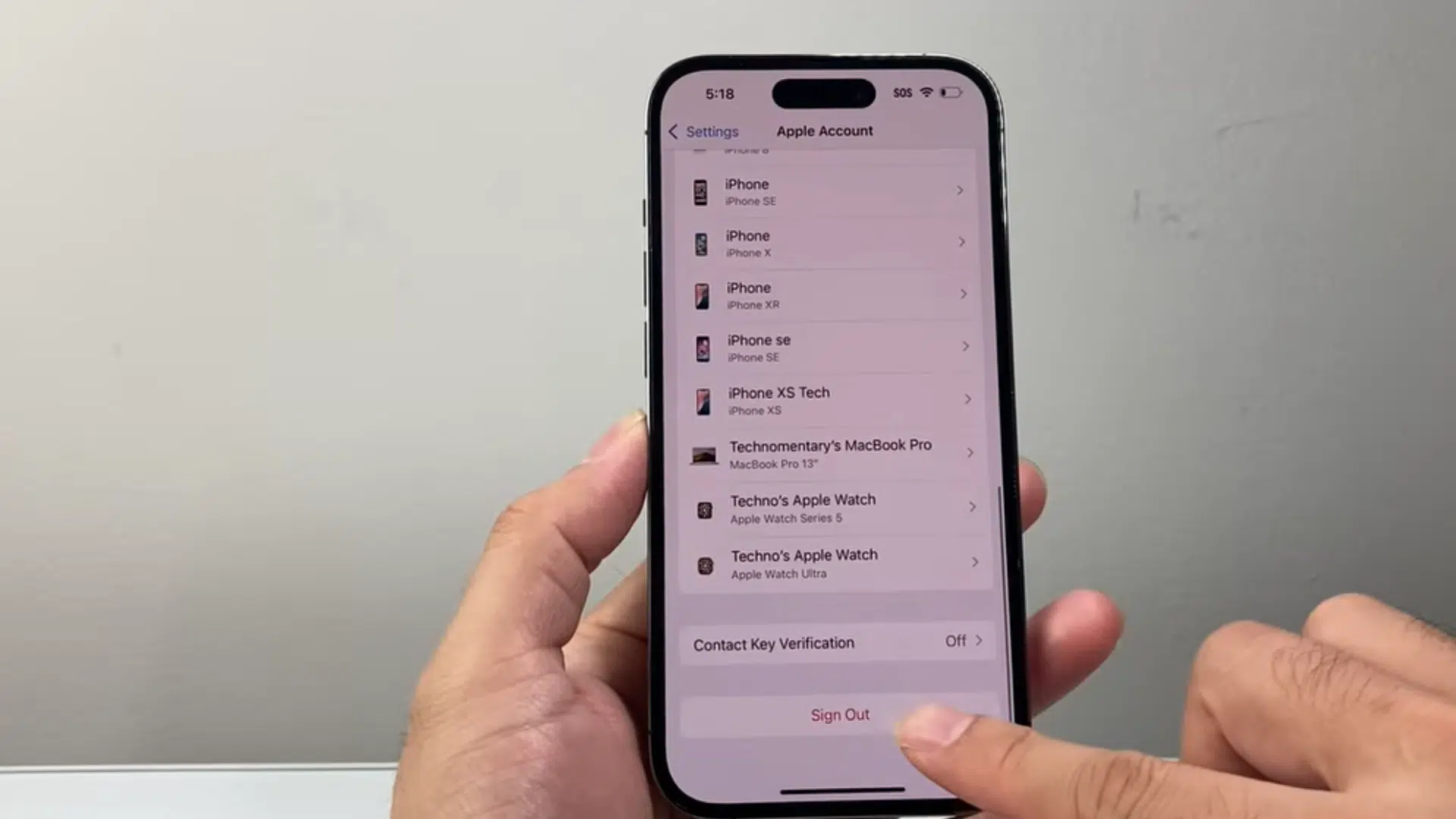 a hand holding an iphone showing apple account settings while tapping the sign out option at the bottom of the screen