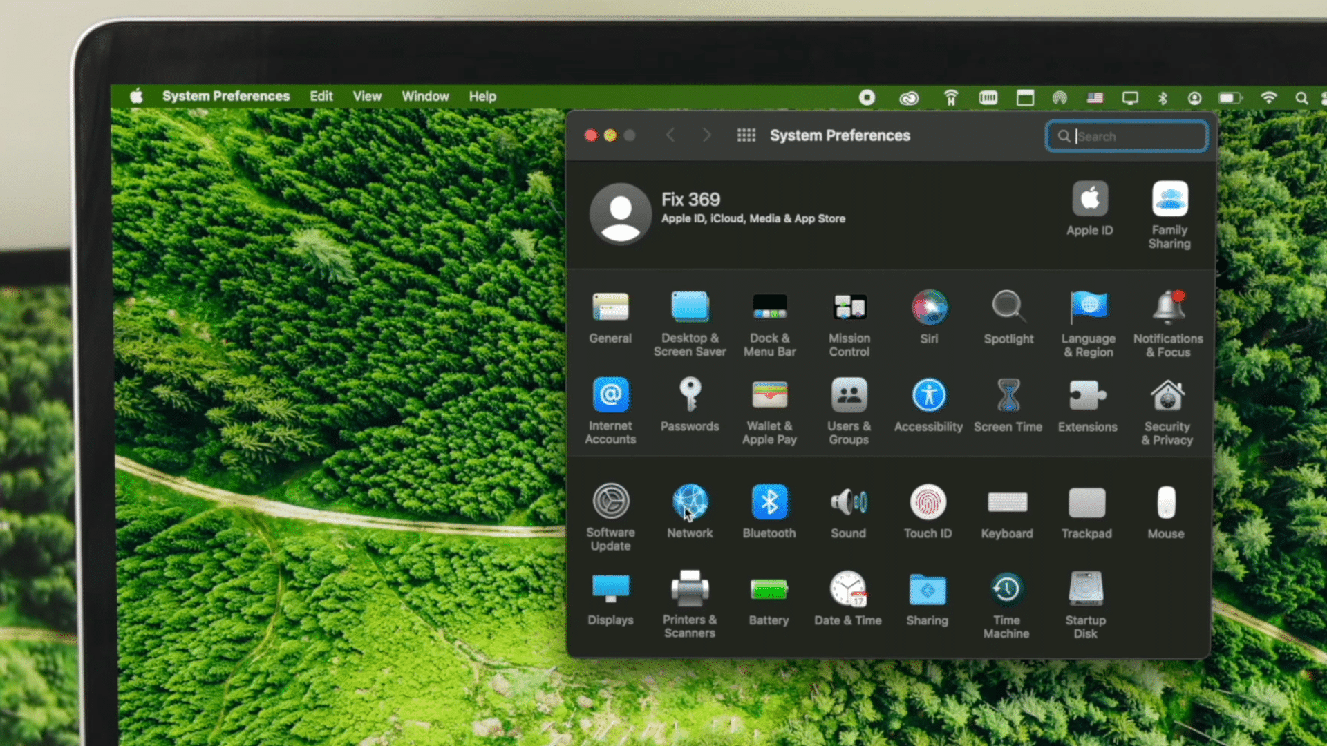 macbook screen showing apple menu opened with system preferences option selected over green forest wallpaper background