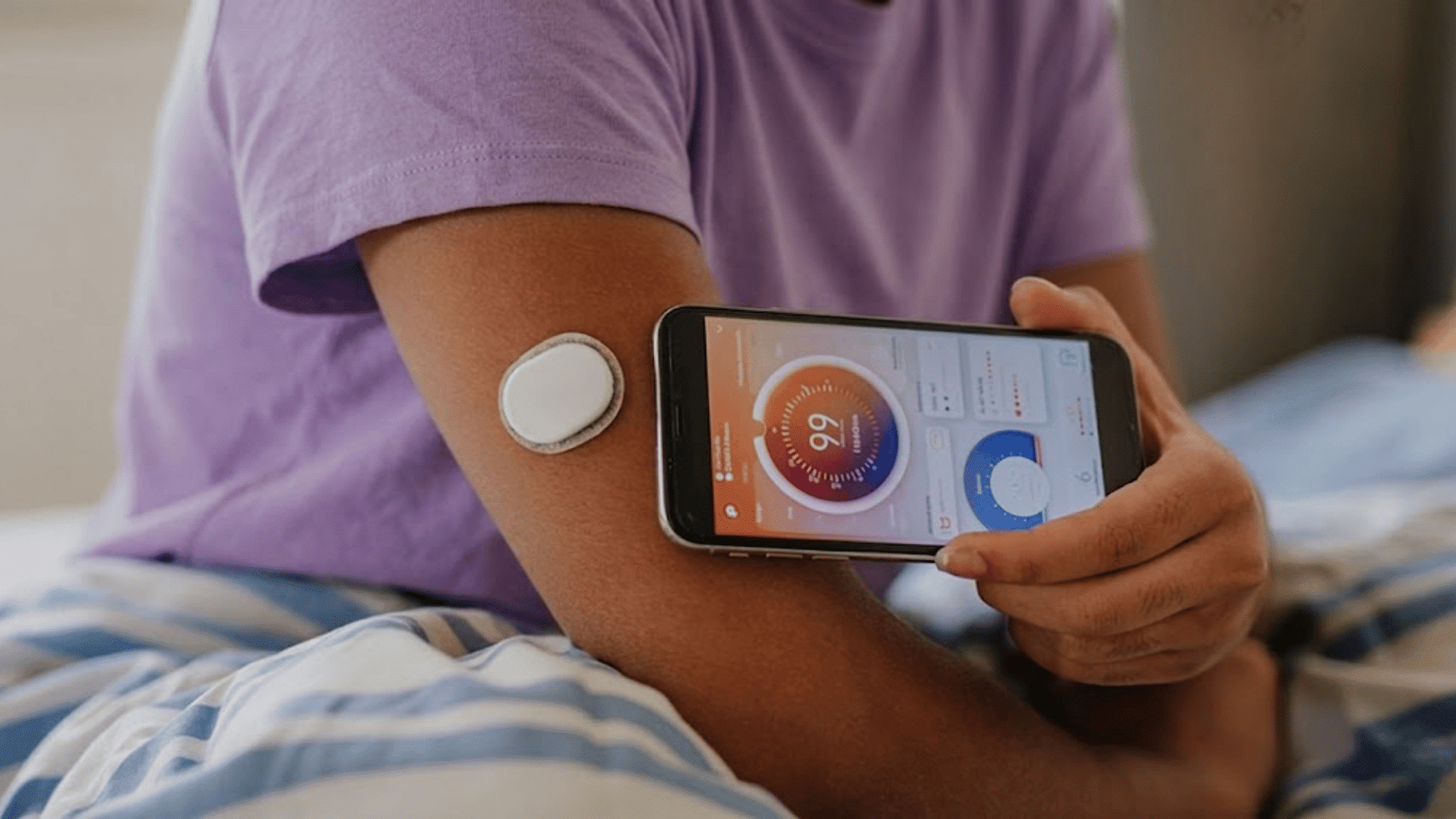 person wearing a glucose sensor on upper arm while holding a smartphone displaying health data in a home setting