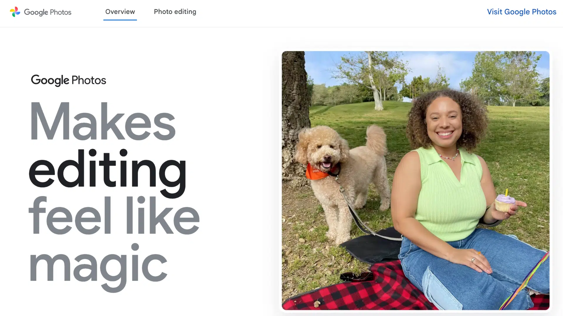 smiling woman holding a cupcake sits on a picnic blanket next to her fluffy dog in a grassy park