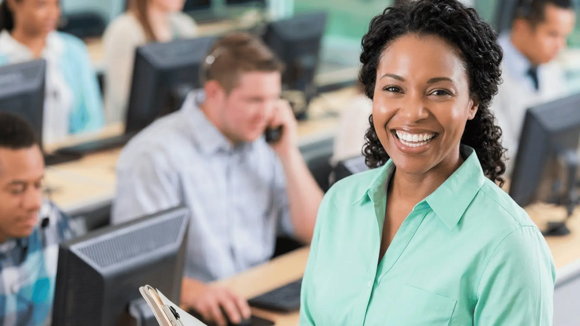 smiling woman in office training room with coworkers using computers in the background