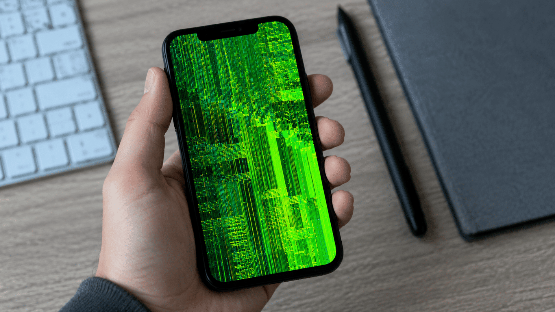 hand holding smartphone with bright green screen glitch displaying digital distortion over desk workspace background