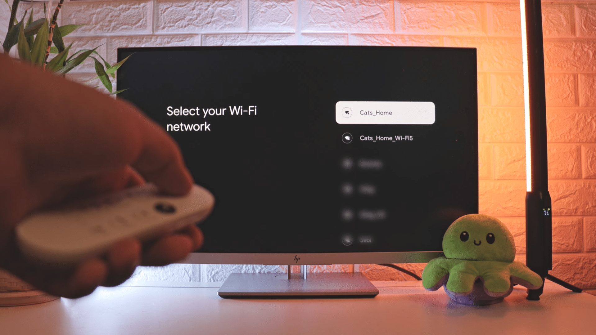 hand using a remote control to select a wifi network on a television setup screen with a plant plush toy and ambient lighting in the room