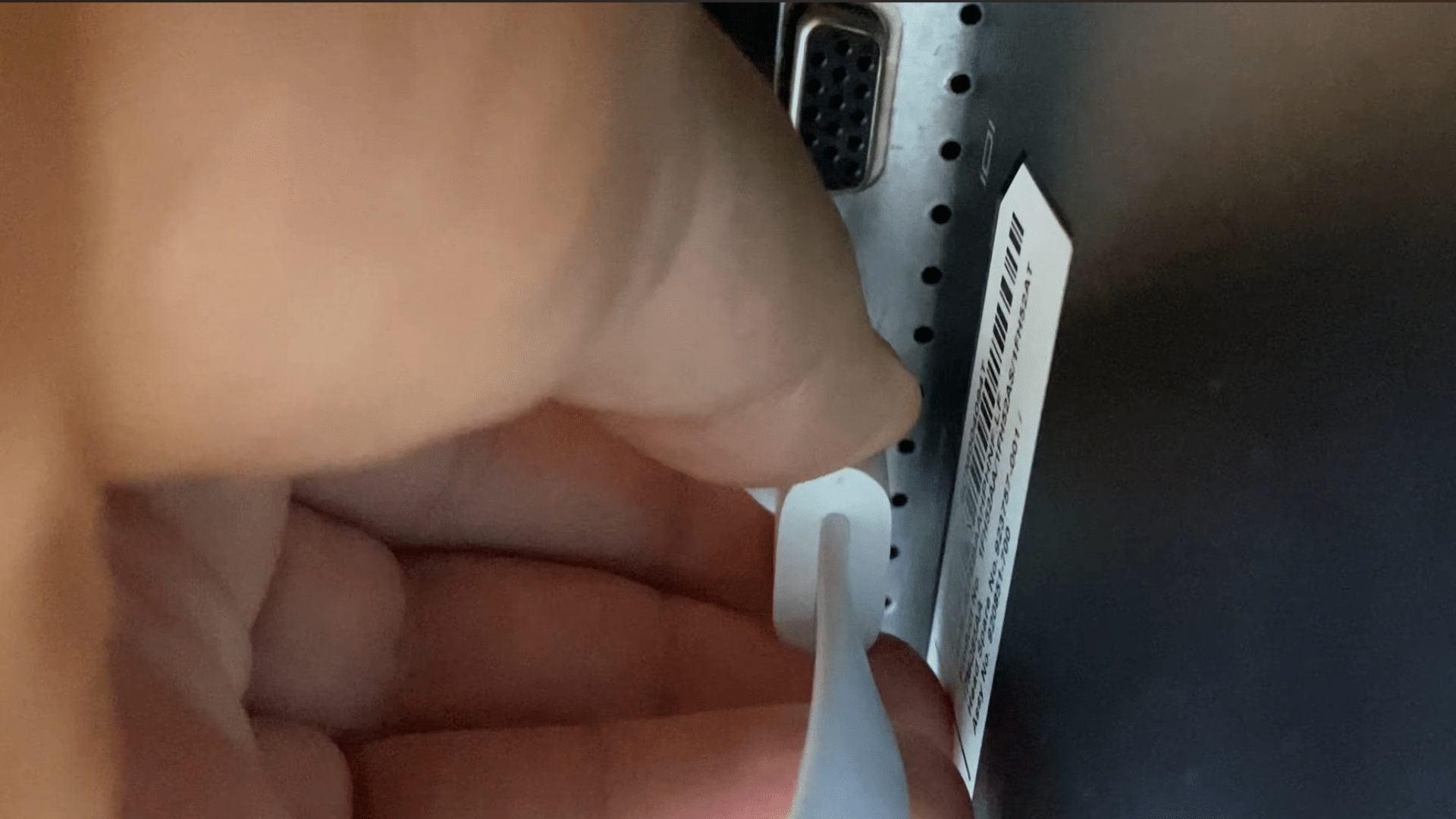 hand using a white tool to press a recessed button on the back of a monitor near a barcode label and port connector