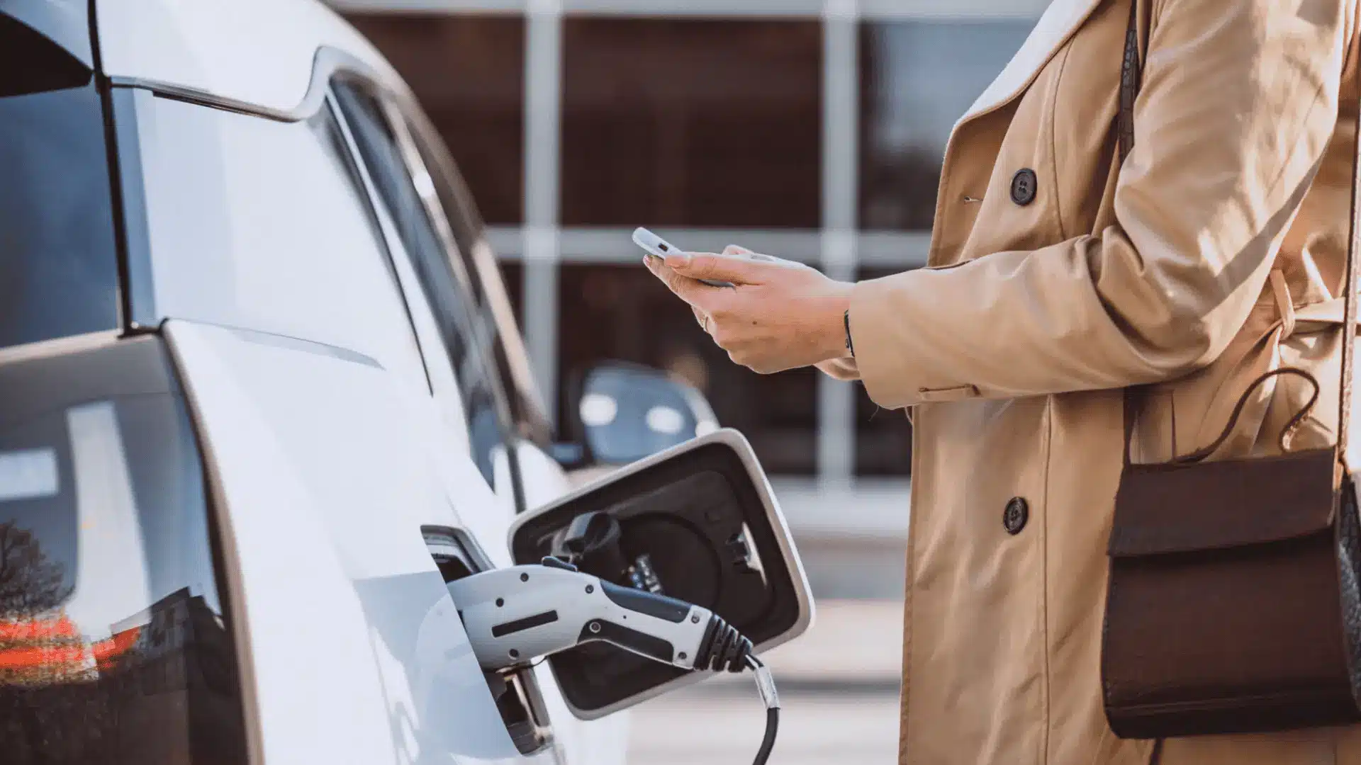 white electric car plugged into a charging cable while a person stands beside it using a smartphone