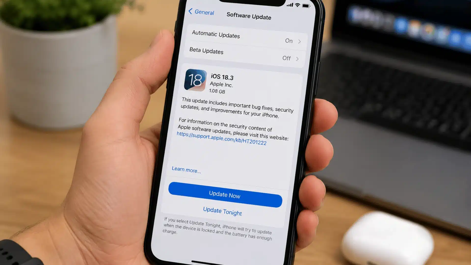 hand holding an iphone on the software update screen beside a laptop plant and airpods case on a wooden desk
