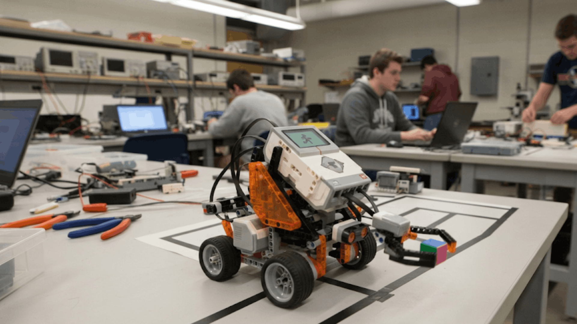 small educational robot vehicle on a testing table in a robotics lab with students working on projects in background