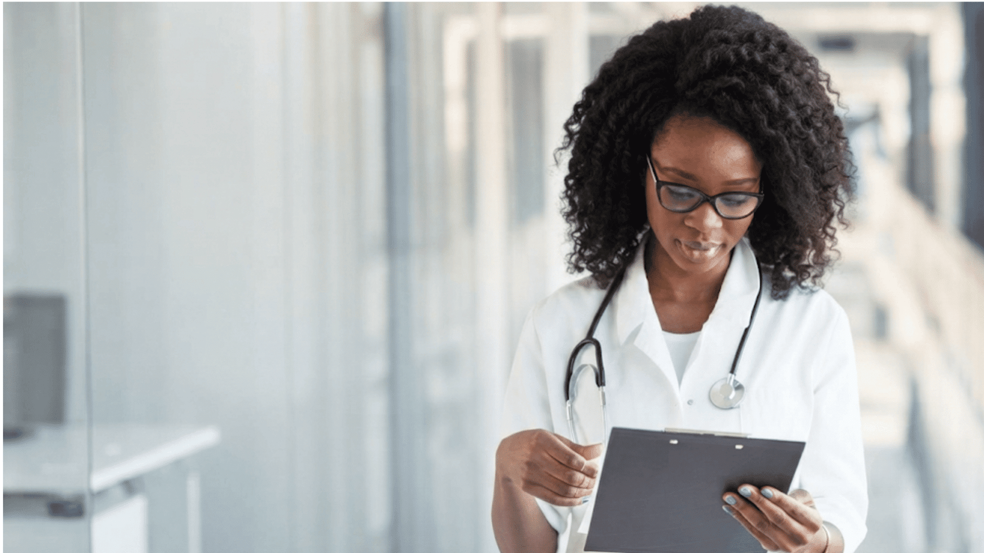 doctor wearing a white coat and stethoscope reviewing information on a clipboard while standing in a hospital hallway