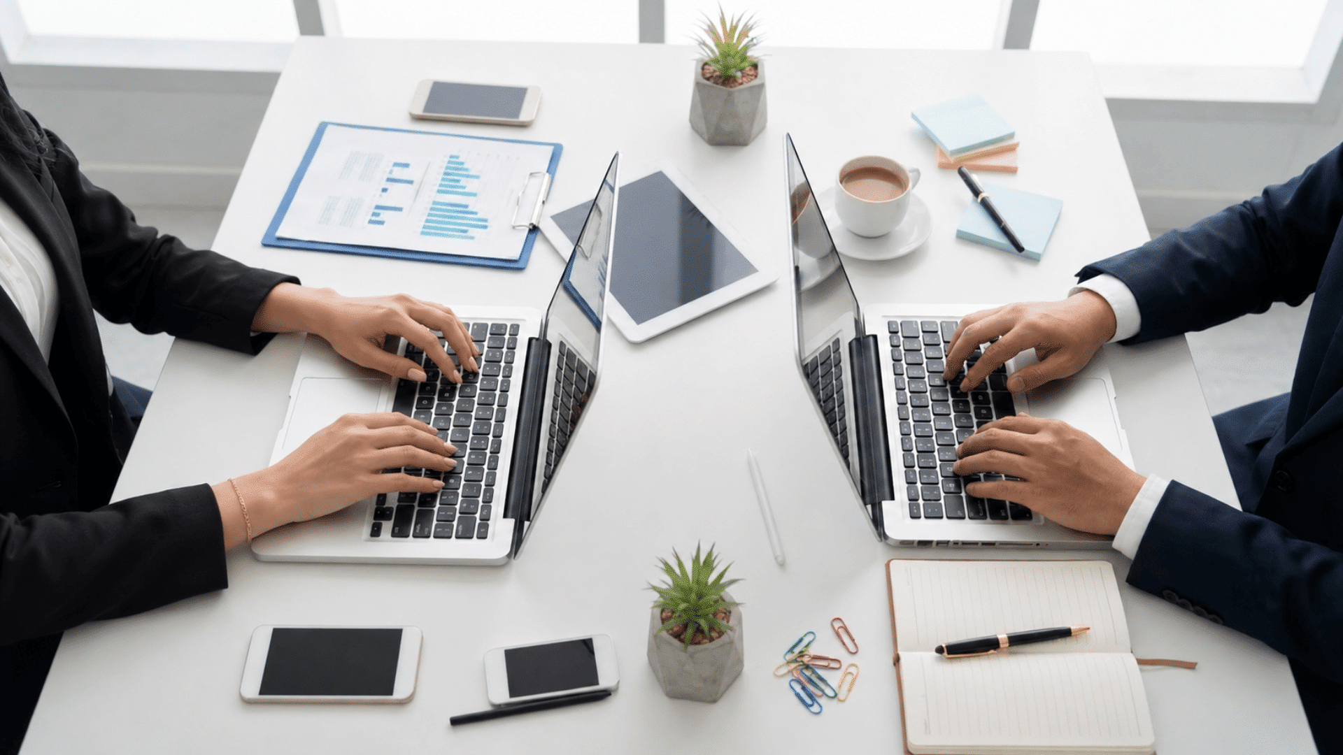 two professionals work on laptops across a desk with charts, tablets, notes, and office supplies.