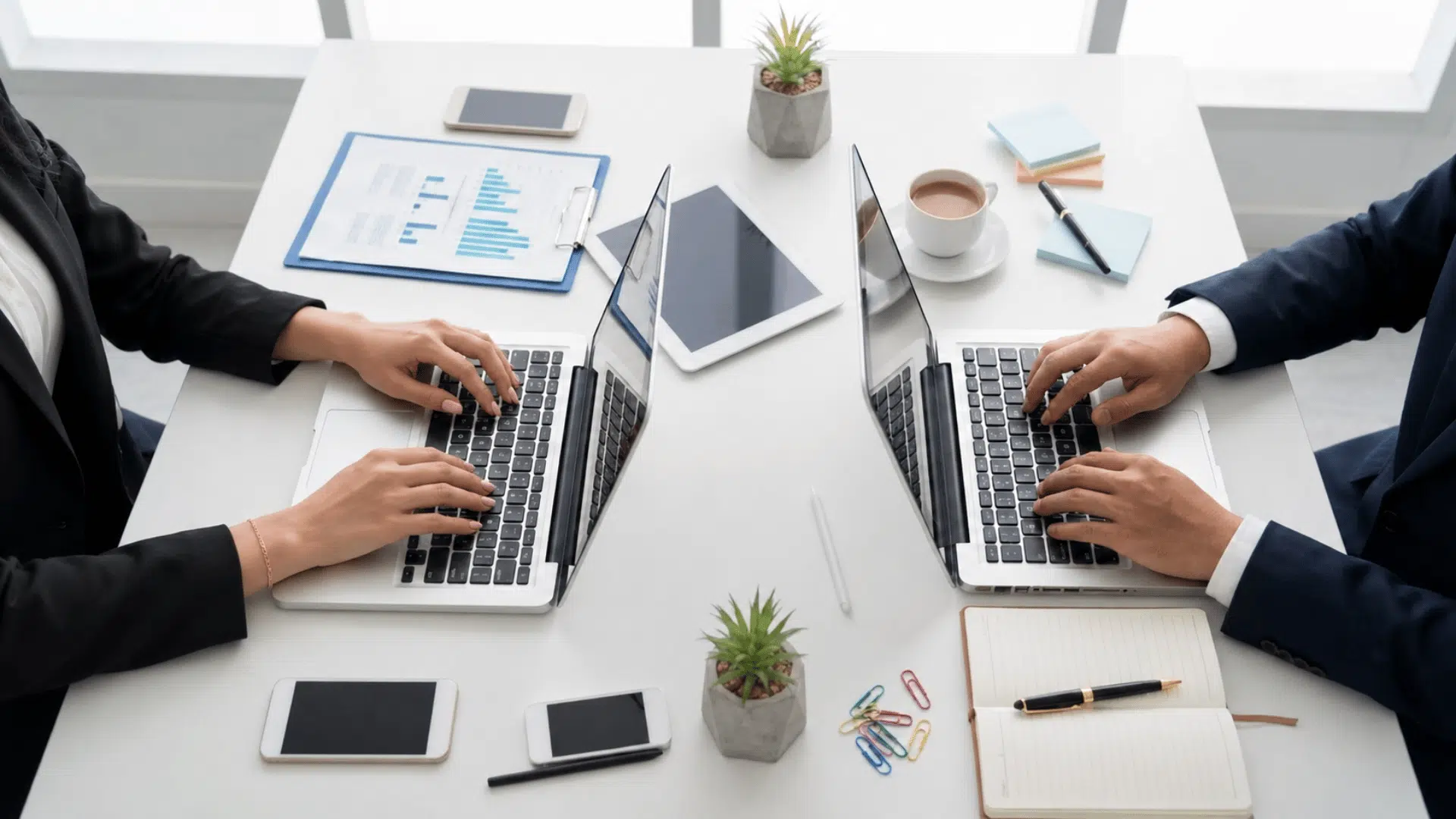 two professionals work on laptops across a desk with charts, tablets, notes, and office supplies.