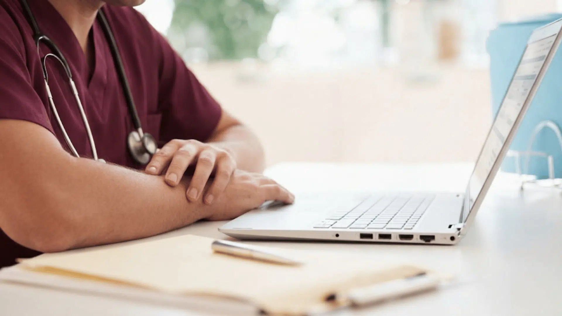 doctor wearing scrubs and stethoscope sits at a desk using a laptop with documents and pen nearby